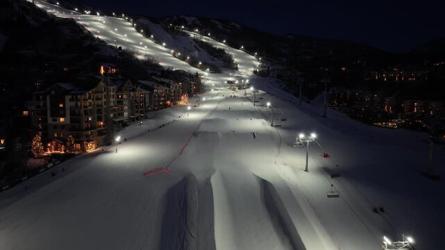 Drone footage resembling Breckenridge, Colorado captures night skiing around a large half pipe, with bright floodlit trails, chairlifts, glowing lodges, and snow covered mountain terrain.