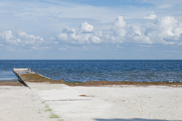 A concrete pier in St. Petersburg, FL stretches from a white sandy beach into a calm blue bay beneath a sky dotted with puffy clouds, suggesting coastal tranquility, wide open horizons, and quiet soli © Del Harper