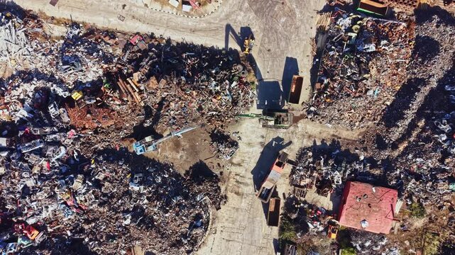 A recycling yard displays large heaps of scrap metal and discarded machinery. Workers operate machinery to sort through materials. The scene is busy with activity under bright daylight.