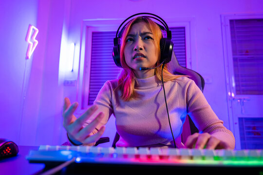 Game over. Angry Asian gamer wearing gaming headphones playing video game on computer PC with purple light on night at home, female tired feeling sad disappointed about game losing