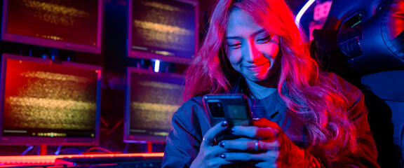 Asian woman in front of a computer monitor display using mobile phone, neon light, Hacker wear black jacket at system control room, Security guard working on multiple monitors together © sorapop
