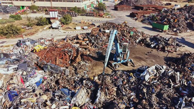 A large area filled with piles of scrap metal and old vehicles is visible. Heavy machinery works on sorting and processing the metal waste under a clear sky.