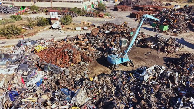 Heavy machinery is working at a scrap yard collecting and sorting metal debris. Various types of metal waste are stacked around the area during a sunny day.