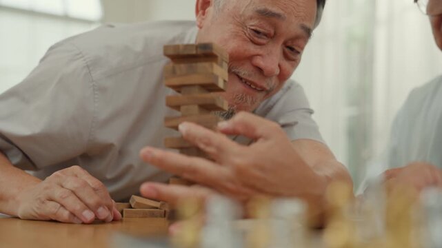 Senior man laugh while enjoying game with wooden block at home wearing casual shirt near bright window, caregiver support, focused play, joyful leisure activity indoor lifestyle