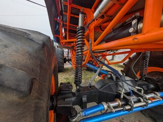 Underneath view of a custom monster truck suspension system featuring heavy-duty coilover shocks, bright orange tubular chassis frame, hydraulic steering rams, and massive off-road tires. © Kevin