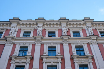 Riga, Latvia – View of classical building facade from street level, showing caryatid and telamon stone sculptures supporting upper cornice above symmetrically arranged dark window frames on wall. © Mltz