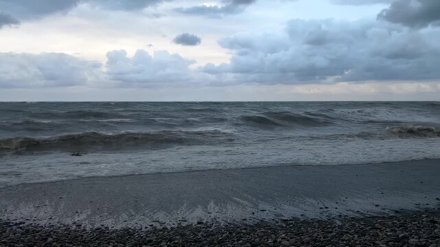 Wide shot of waves breaking on pebble beach, with rough Black Sea surf rolling toward the Batumi shoreline under heavy clouds, useful for travel, weather, coastal tourism and environmental projects