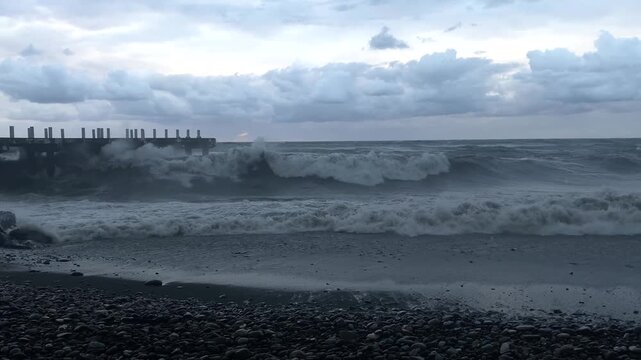 Wide shot of storm waves hitting a coastal pier, with white surf washing over the rocky Black Sea shoreline in Batumi, Georgia, for weather coverage, travel campaigns and coastal environment stories