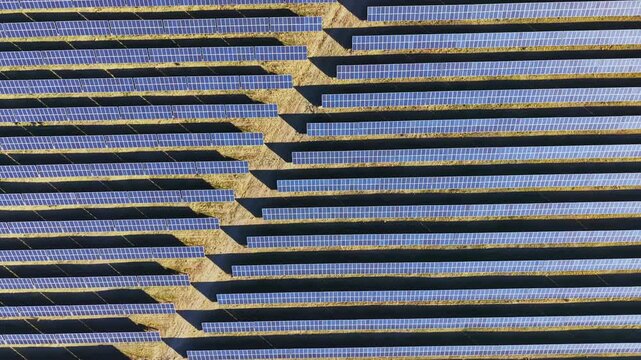 Solar panels cover a large area arranged in parallel rows. The scene shows a clear sky above, with sunlight reflecting off the panels.