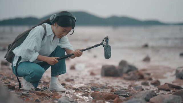 Young woman wearing headphones and recording ambient nature sound with a boom microphone on a muddy tidal flat