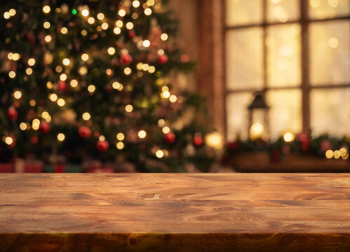 Rustic wooden table stands beneath glowing Christmas lights, with decorated evergreen tree and bright window softly out of focus. Warm amber bokeh gives festive interior welcoming mood.