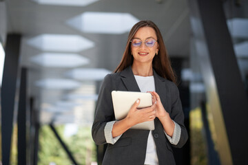 Professional woman uses tablet with contented smile, reflecting technology adoption in business. Tablet user touches screen while standing in contemporary office, dressed in gray blazer. © Agil