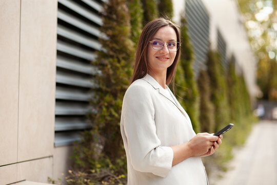 Smiling woman holds smartphone outdoors, glancing aside with relaxed expression. Phone user stands beside modern building, suggesting digital networking and convenient mobile communication.