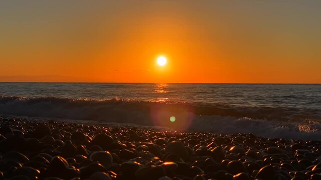 An intense orange sunset lights the Black Sea and its moving waves, with rounded shore stones in the foreground. The uninterrupted horizon makes this coastal footage useful for travel and relaxation