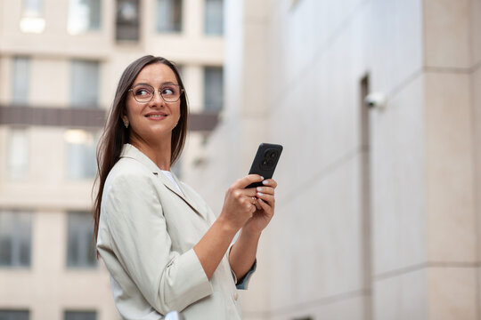 Businesswoman checks smartphone outside office building, smiling as she looks away from screen. Professional in glasses holds her phone with both hands, ready for work conversation.