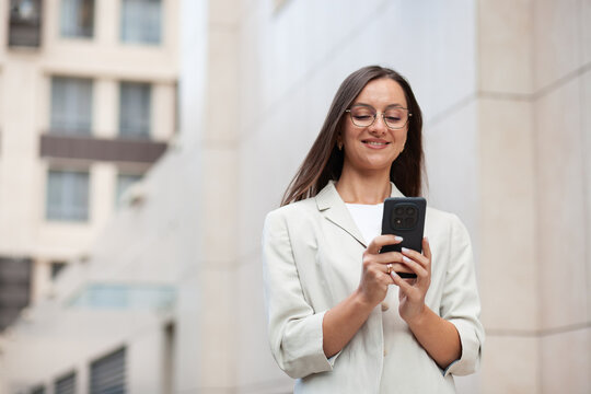 Focused woman uses smartphone outdoors, smiling at what appears on screen. Businesswoman holds her phone at chest level, illustrating digital distraction in bright modern city setting.