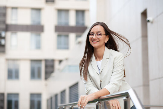 Smiling businesswoman leans on railing outside modern office building. Professional woman wears glasses and pale blazer, presenting confident, approachable image suited to mobile banking services.