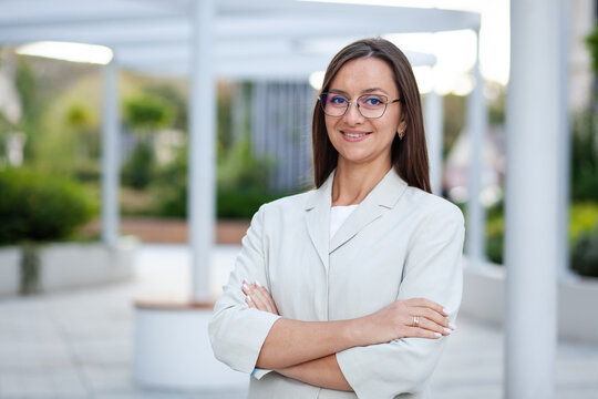 Smiling businesswoman poses with arms folded in landscaped office courtyard. Confident executive in glasses and light blazer presents polished, approachable image of professional leadership.