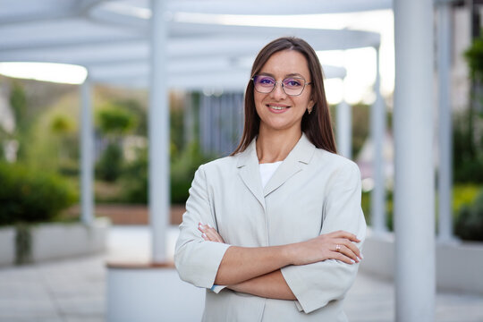 Woman in light blazer smiles with arms crossed outside contemporary workplace. Her polished career portrait, framed by glasses and clean glass architecture, conveys confidence and approachability.