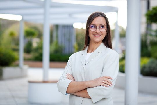 Smiling businesswoman poses with folded arms in bright office courtyard, conveying professional confidence. Woman in eyeglasses and light blazer looks composed among landscaped greenery.