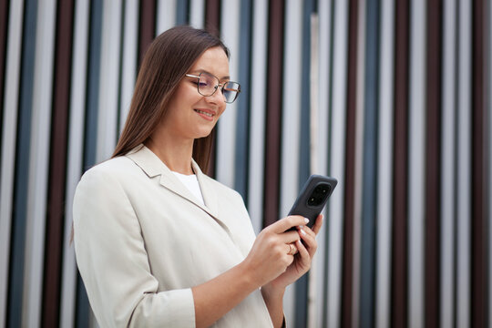 Woman smiles while checking smartphone outdoors, holding device in both hands. Eyeglass wearing professional focuses on mobile screen against contemporary facade of tall alternating vertical panels.