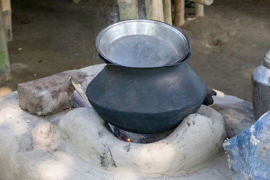 Cooking pot placed on a traditional mud chulha stove with steam rising. Rural South Asian outdoor kitchen preparing food over wood fire flame.