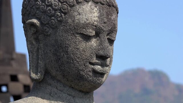 Stone Buddha statue at Borobudur temple with ancient stupas and blue sky background in Central Java Indonesia.