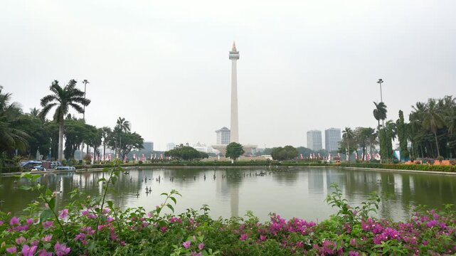 Jakarta, Indonesia - October 12, 2026: Wide view of the National Monument (Monas) rising above a reflecting pond, with landscaped gardens, flowering plants, palm trees, and city buildings in the backg