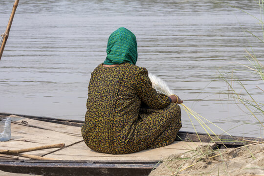 Rear view of a South Asian woman in hijab sitting on wooden boat holding wild grass flower near river. Autumn rural peace concept.