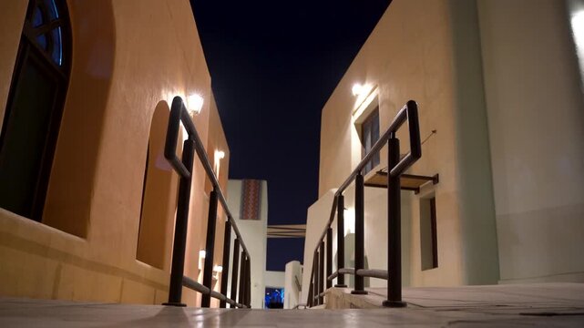 Low angle view walking down stone stairs in a narrow, illuminated alleyway at night, showcasing traditional middle eastern architecture with mud brick buildings and dark wooden handrails