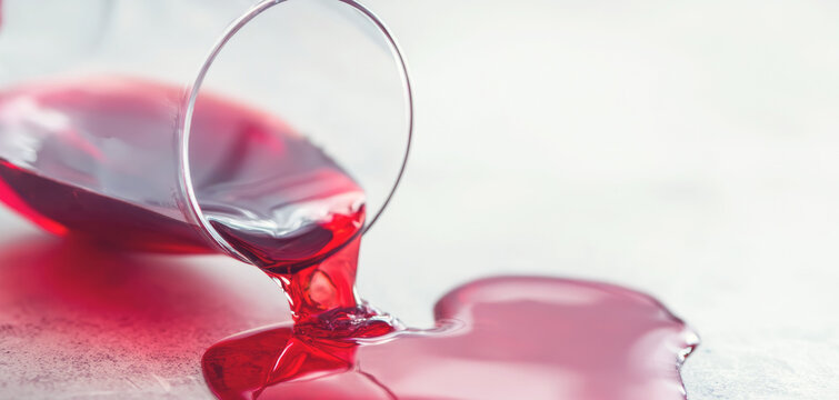 Red wine spilling from a tipped over glass, forming a spreading puddle on a light colored floor, indicating an accident or mishap