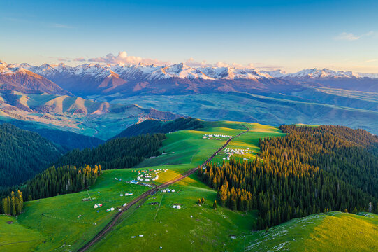 Aerial view of golden sunset light illuminating snow-capped mountain peaks and green forests in Kalajun Grassland, Xinjiang, China.