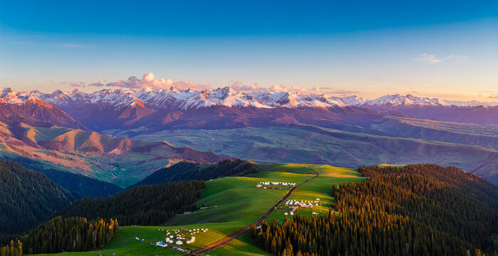 Aerial view of golden sunset light illuminating snow-capped mountain peaks and green forests in Kalajun Grassland, Xinjiang, China.