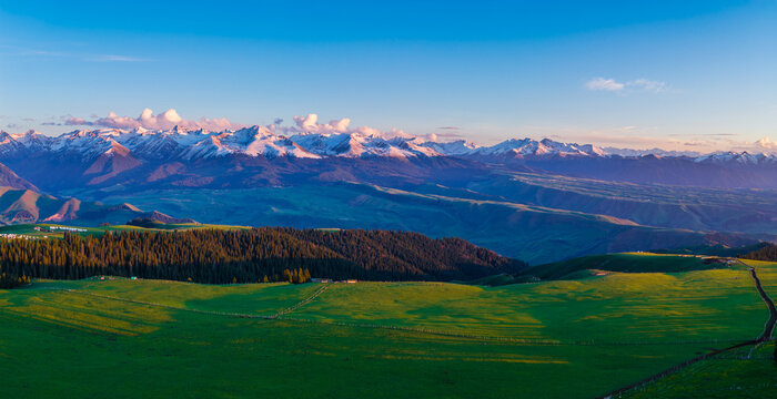 Aerial view of golden sunset light illuminating snow-capped mountain peaks and green forests in Kalajun Grassland, Xinjiang, China.