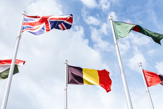 United Kingdom Belgium and partially visible flags waving on white poles against a bright blue sky with clouds in an outdoor international display