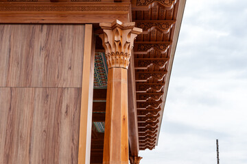 Ornate carved wooden architectural facade with column, decorative eaves, geometric details, and cloudy sky beside a traditional building exterior © tashmetova808