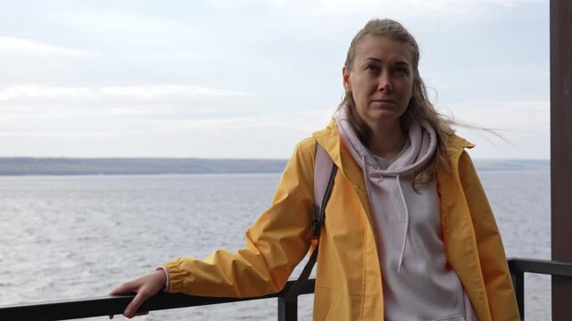 Thoughtful young woman in a yellow jacket standing on a balcony with her hair blowing in the wind, looking pensive and sad against a vast sea and cloudy sky background on a cool day