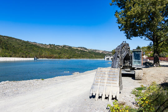 A large tracked excavator parked on a dirt path next to a serene river and dam structure under a bright blue sky.