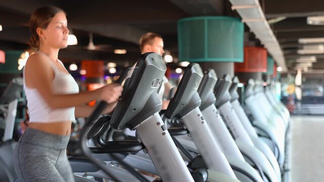 Young athletic woman in sportswear training on elliptical machine in gym 
