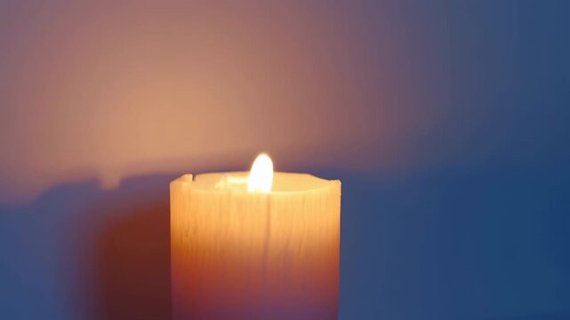 Pillar candle burns with flickering flame above textured yellow wax, casting shifting amber light and soft shadows against blue background in close-up with generous copy space