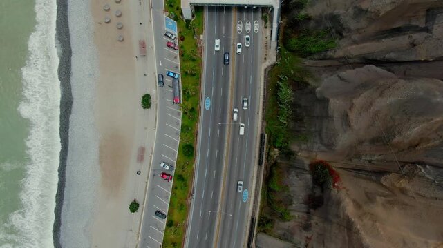 Overhead drone glides forward over the Costa Verde in Lima, Peru, following a pedestrian bridge, traffic lanes, beach umbrellas, surf and the rocky cliff in parallel strips.
