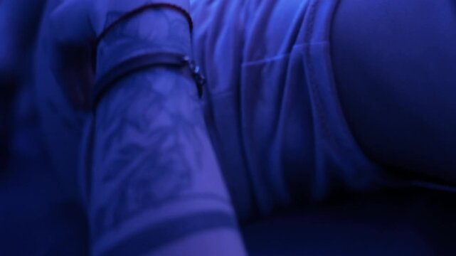 Close-up shot of hand resting and lightly pressing on upper thigh wearing patterned lace lingerie and underwear, wrist adorned with bracelets and forearm tattoo visible