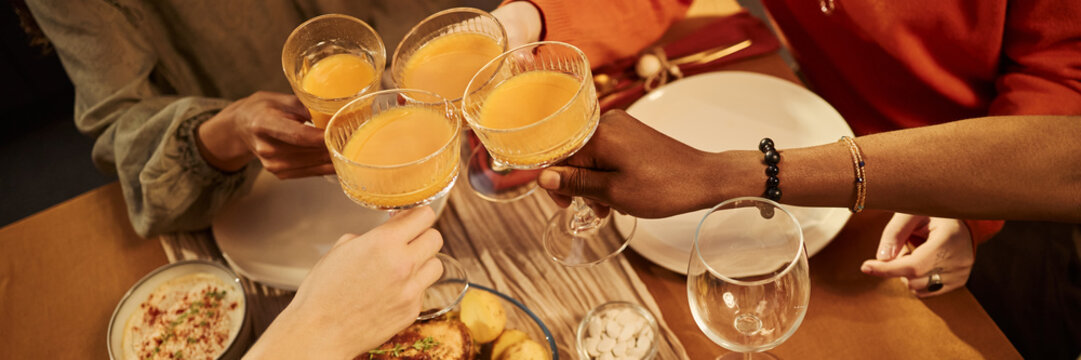 Header of African American and Caucasian young adults toasting cocktail glasses over dinner table during festive celebration. Image suitable for beverage advertising campaigns
