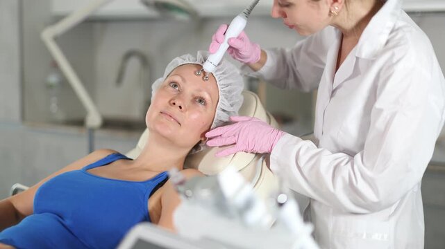 Adult woman cosmetologist performs hardware facial microcurrent procedure to young female patient 