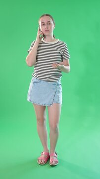 Portrait of elegant teen girl dressed in summer T-shirt and light blue jeans skirt talking by smartphone standing against green screen 