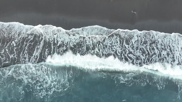 Ocean waves crash onto dark sandy beach, creating foam and ripples, with a clear blue water gradient visible in the foreground and rocky shoreline in the background. Black volcanic sand. Aerial view