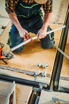 Plumber in overalls kneeling on floor insulation cutting a white ppr pipe with red manual pipe shears during underfloor water supply installation.