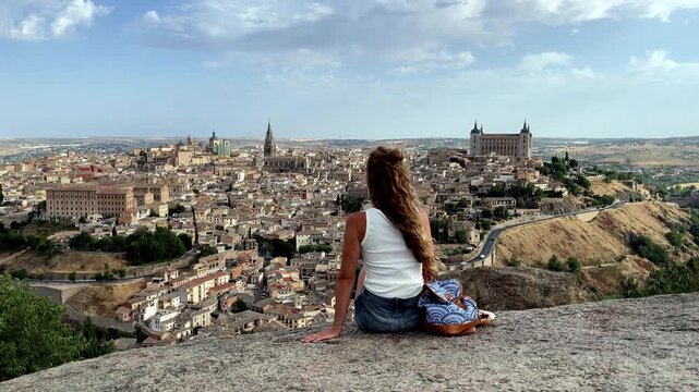 back view of tourist woman sitting on stone wall looking at toledo medieval city skyline and alcazar in castile la mancha spain