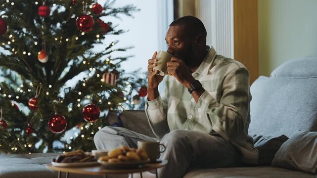 Young man drinking from a ceramic cup while sitting on the sofa beside a decorated Christmas tree, enjoying a quiet holiday break at home