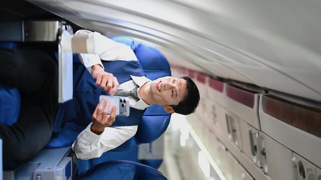 A young Asian executive smiles while using his smartphone in business class. Modern corporate travel, mobile connectivity, and remote work lifestyle during a flight.